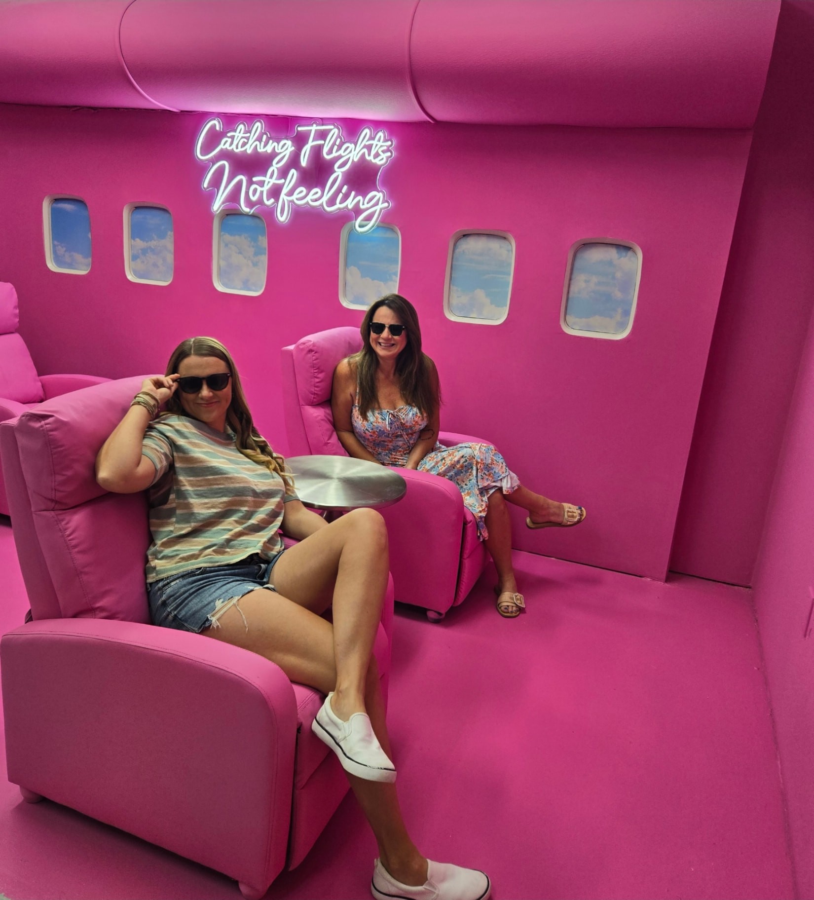 Guests relaxing in pink recliners in the private jet room with a Catching Flights Not Feelings neon sign